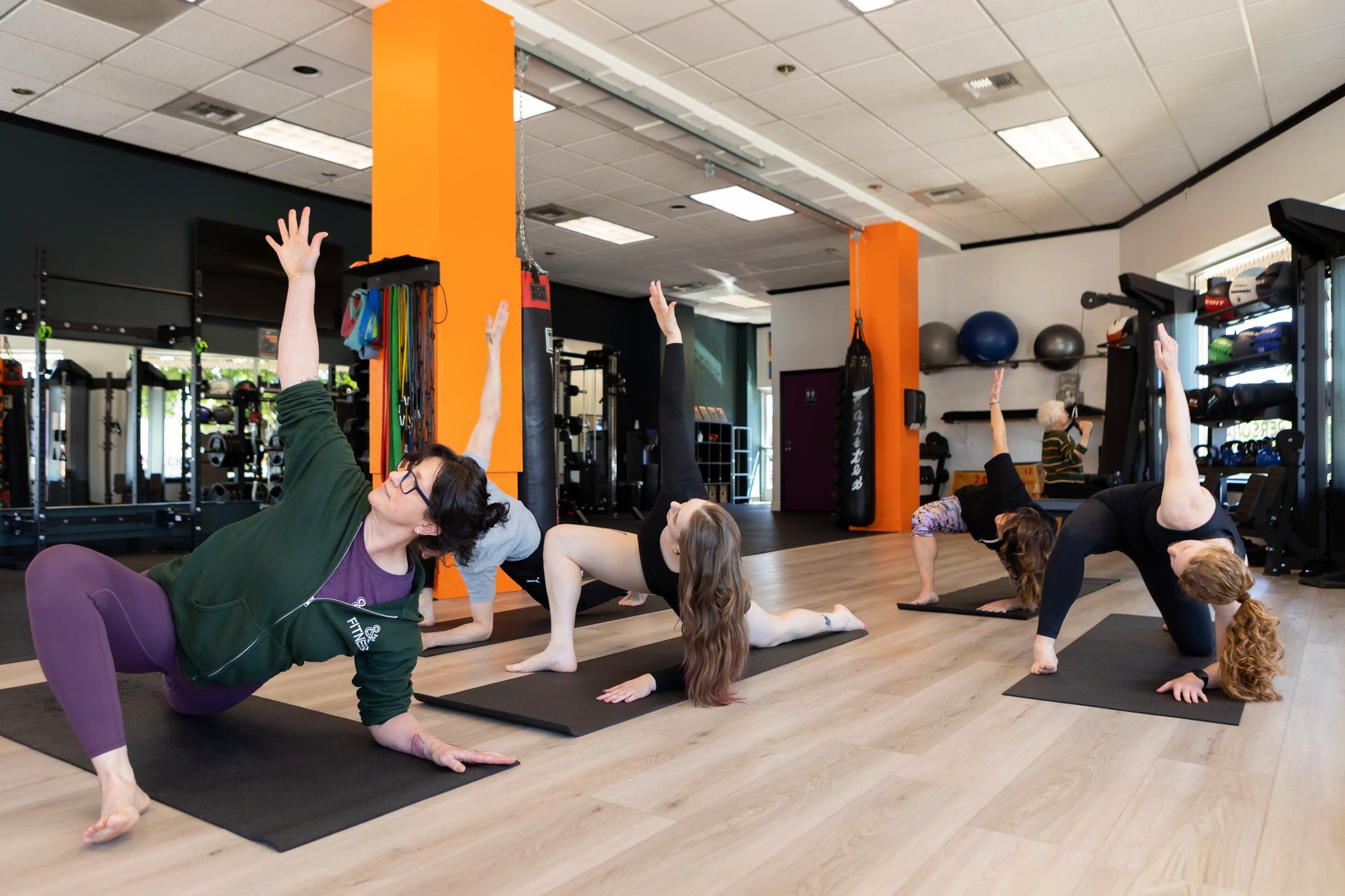 A group fitness/stretch class in a gym: five people on black mats in a side-plank/stretch pose, one in a dark green hoodie with a visible '& FITNESS' logo, reaching upward together; the gym floor behind them shows racks, resistance bands, a punching bag, an orange support column, and an older adult using equipment in the background.