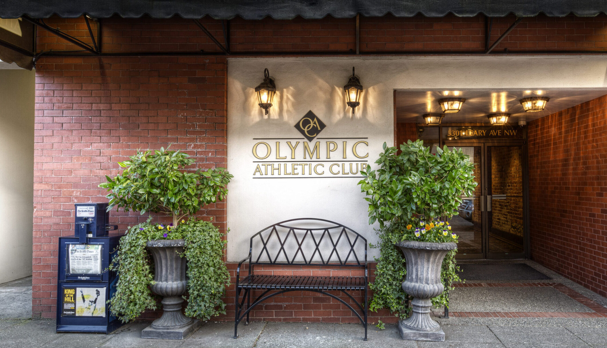 The Olympic Athletic Club entrance on Leary Avenue in Ballard: red brick, a cream signboard reading OLYMPIC ATHLETIC CLUB with a diamond monogram, carriage lamps, and 5301 LEARY AVE NW lettered above the doorway.