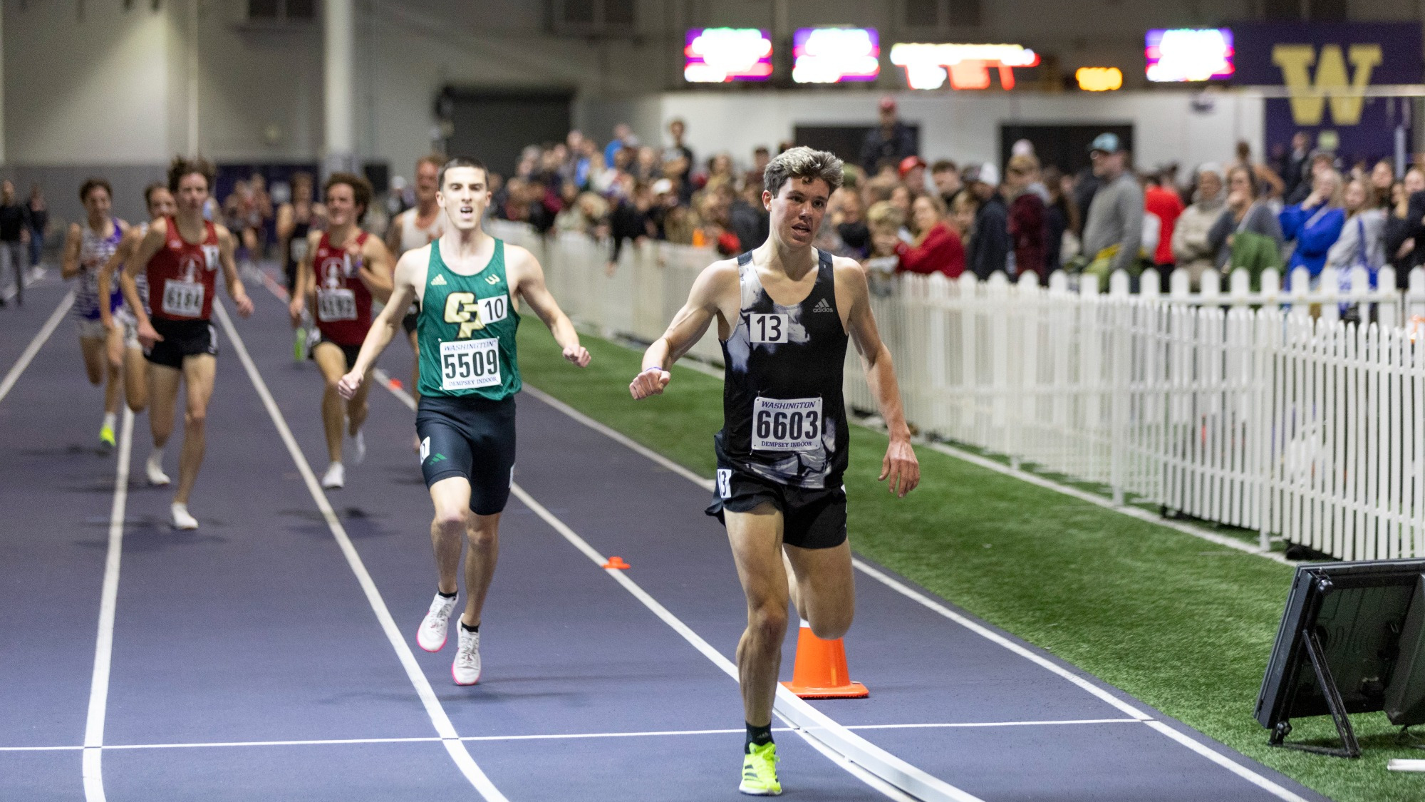 Six runners mid-race on the purple oval inside the Dempsey Indoor, bibs reading Washington Dempsey Indoor, a standing crowd behind a white picket fence and a gold W on the far wall.