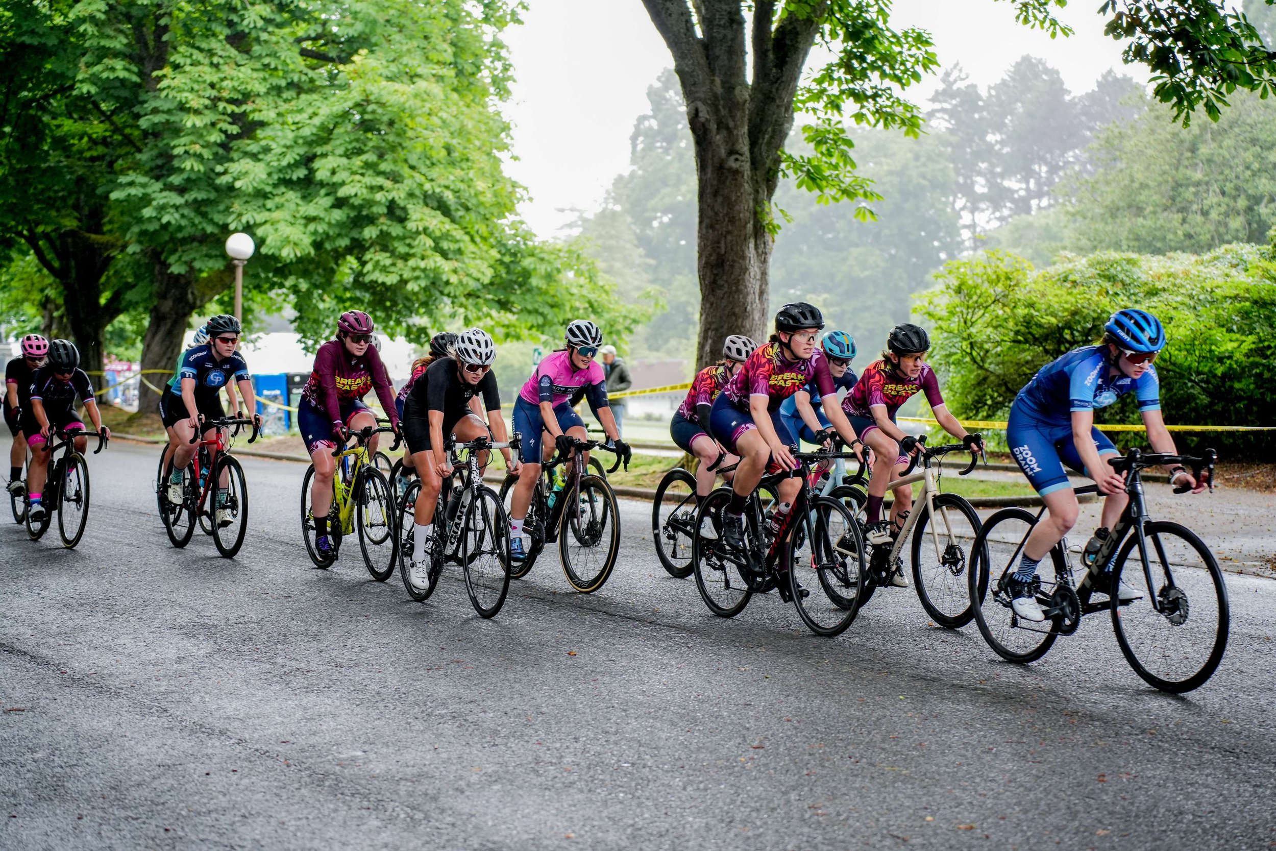 The pack mid-race on a wet, tree-lined park road, team kit through the middle, course tape behind.