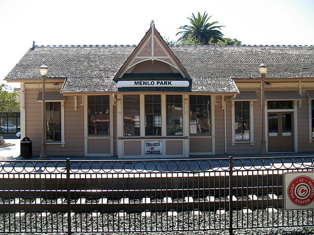 Historic Menlo Park train station with wooden facade, black roof, palm tree in background; sign says "MENLO PARK." Calm atmosphere.