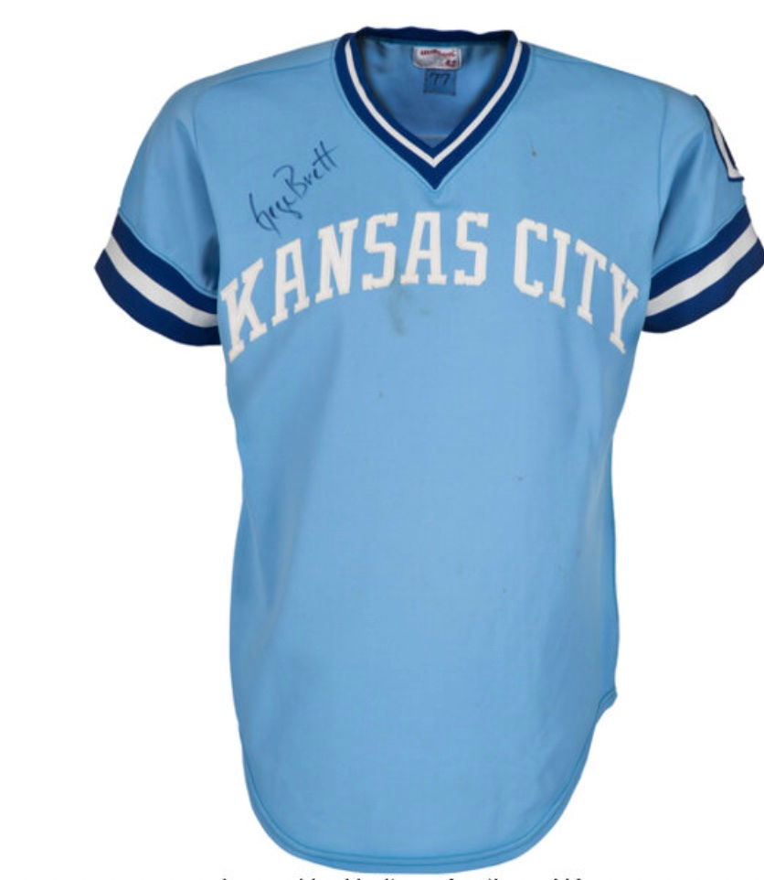Light blue Kansas City Royals jersey from 1977, manufactured by Wilson, featuring white lettering, blue and white striped sleeves, and George Brett’s autograph on the upper left chest.