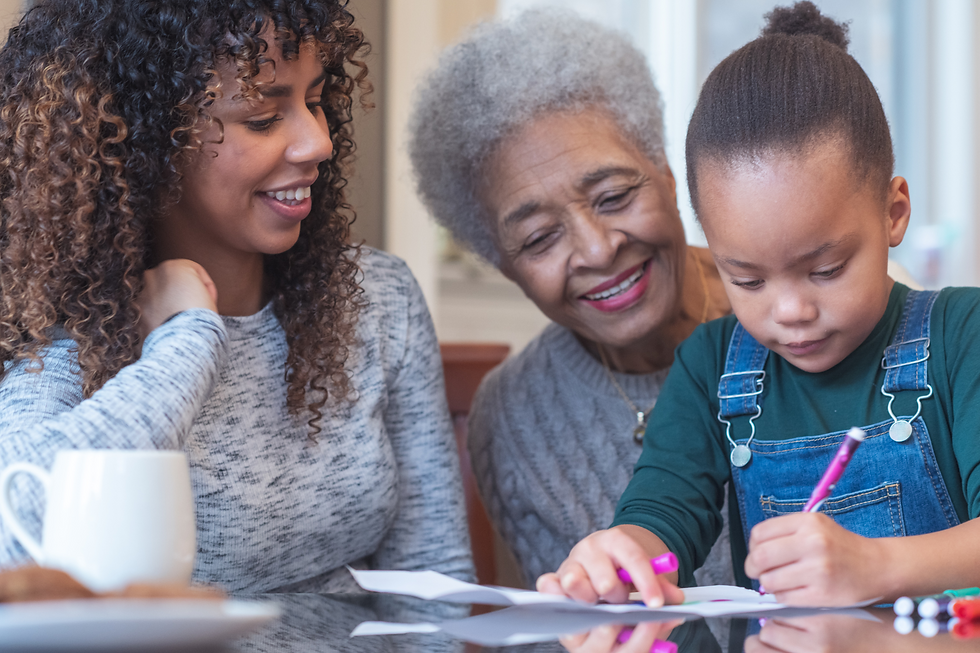 Three generations of women—grandmother, mother, and daughter—sitting together at a table, with the young girl drawing while the grandmother and mother watch with smiles, highlighting familial love and support.