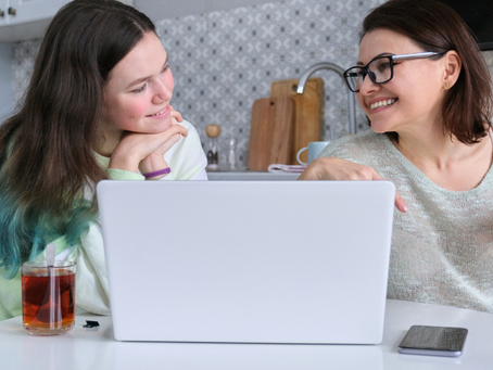 Two women smiling and talking while looking at a laptop together at a kitchen table.