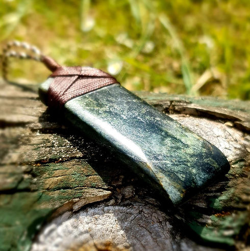 Hand-carved West Coast Serpentine pendant resting on weathered wood, showing deep green and golden tones with a natural braided cord.
