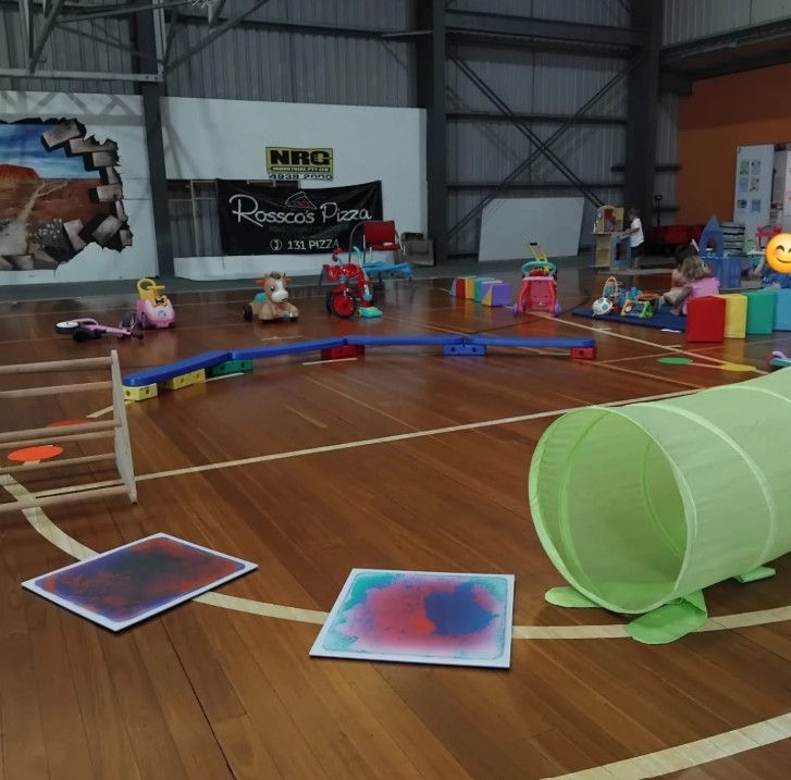 Indoor space with colourful obstacle course, toy ride ons and tunnels. there are some children playing in a soft play space.