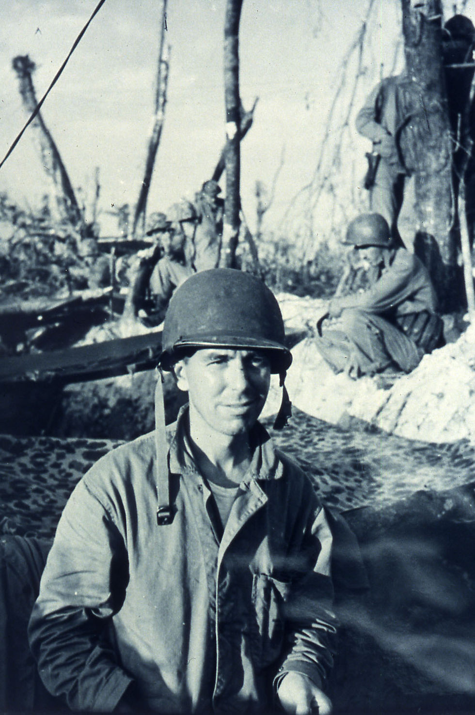 Soldiers in helmets rest in a barren landscape with scattered trees. One soldier looks at the camera, conveying a somber mood. Black and white.