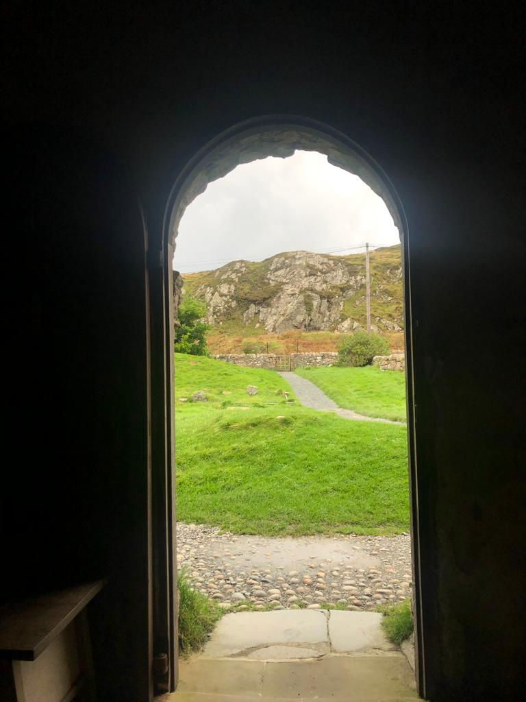 Doorway from dark chapel looking out on to green grass and rocky mountain beyond