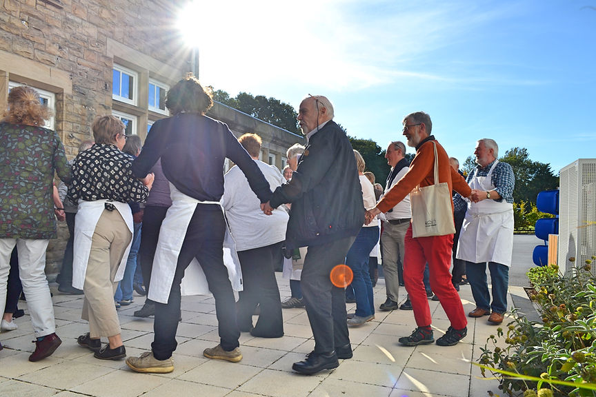 A group of adults holding hands to create a chain  and moving in a spiral.  They are outside  in a court yard with a stone built building to the side, the sun is shining overhead