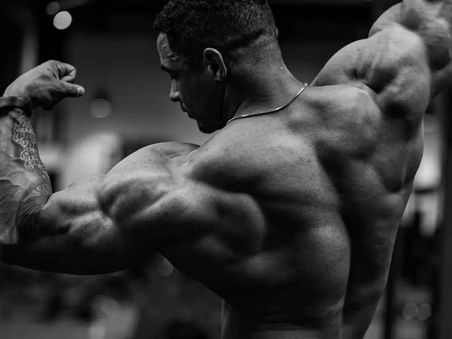 Black-and-white image of a muscular bodybuilder flexing his back and arms during an intense gym workout, highlighting strength, muscle definition, and advanced bodybuilding performance related to Buy Ultima-TNE 100 Online.