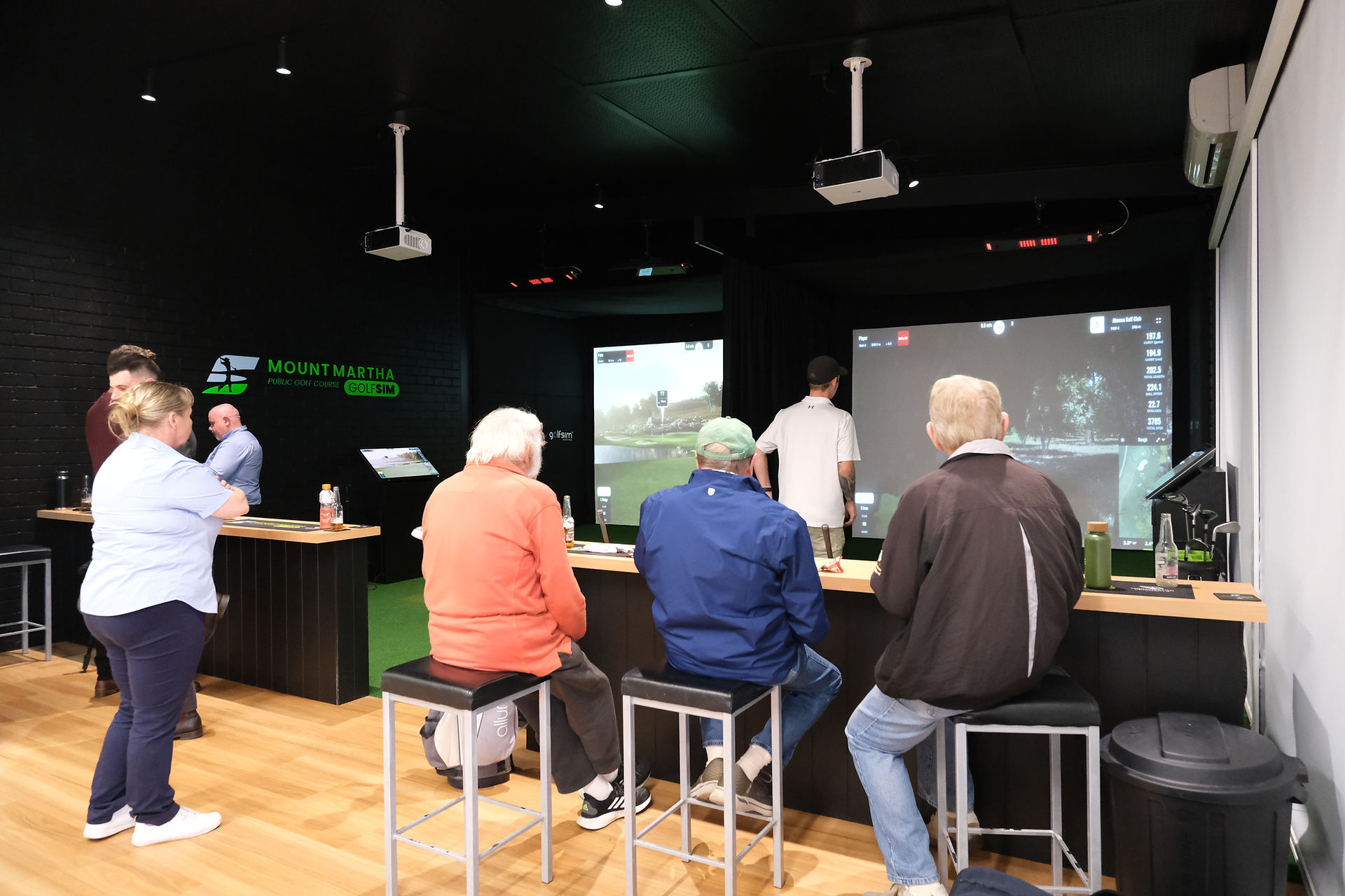 Group of guests seated at bar stools inside the Mt Martha GolfSim room, watching live play on dual GSPro simulator screens during the grand opening event.