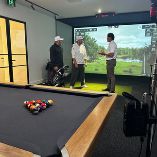 Daisy Thomas and two men discussing golf inside a luxury golf simulator room, with a pool table and golf bags in the background.