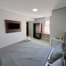 A perspectiva da chegada a um refúgio de descanso, focada na mão de uma viajante segurando a alça de uma mala rígida texturizada. O olhar é guiado para o interior da suíte iluminada pelo sol, onde a cama perfeitamente arrumada exibe toalhas enroladas em tons terrosos sobre o lençol branco, simbolizando as boas-vindas e o início de uma experiência de estadia relaxante.