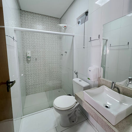 Um banheiro majestoso revestido inteiramente em mármore polido de tons quentes, evocando a atmosfera de um spa privativo exclusivo. Em primeiro plano, uma banheira de imersão branca com design curvilíneo contemporâneo atua como peça central. A profundidade do ambiente revela um box de vidro espaçoso e marcenaria fina, tudo valorizado por um projeto de iluminação cênica que reflete nas superfícies brilhantes.