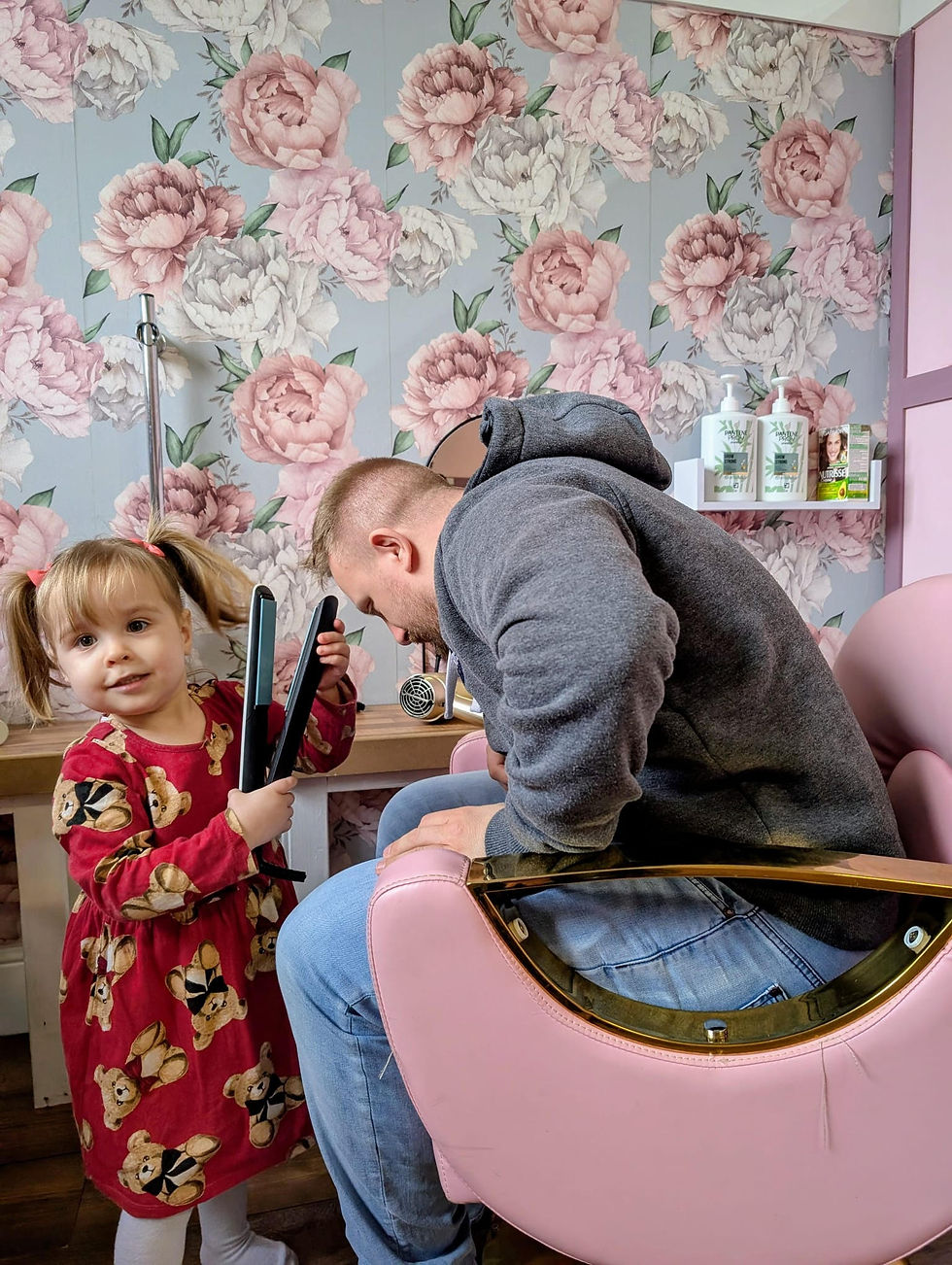A dad sitting in the play hairdressing chair at Kiddos Kingdom Coalbrookdale while his toddler daughter styles his hair with toy straighteners