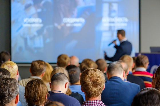 Image illustrative représentant une conférence d'entreprise vue de dos, avec la scène en face, un conférencier micro à la main