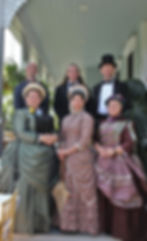 six individuals in Victorian attire grouped on the stairs leading to a porch