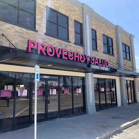 Brick building with "PROVECHO Y SALUD kitchen and bar" sign in pink. Glass doors reflect street scene. Blue sky, bright and sunny.