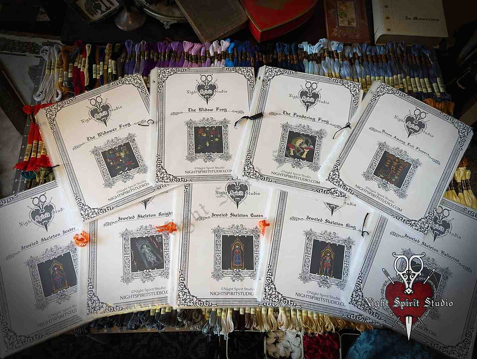 Cross-stitch patterns from Night Spirit Studio arranged on a table, colorful threads in background. Each pattern has ornate borders and dark images.