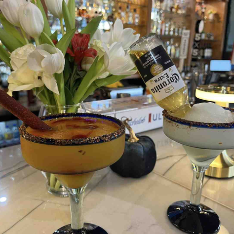 Two cocktails on a bar with a vase of white flowers. One drink has a bottle of Corona, the other a tamarind stick. Bright, lively setting.