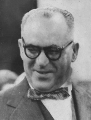 Retrato em preto e branco de João Guimarães Rosa nos anos 1960, usando óculos, terno e gravata borboleta, com leve sorriso.