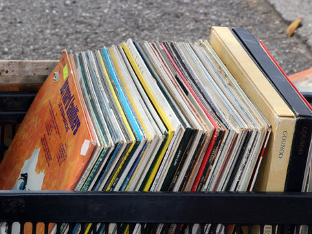 **Texto alternativo (alt text):**  Uma caixa de plástico preta vazada cheia de discos de vinil enfileirados, vistos de um ângulo levemente superior. Os discos estão inclinados para a esquerda. O primeiro vinil, posicionado na frente, possui uma capa predominantemente laranja. Ao longo da caixa, veem-se diversas lombadas finas e coloridas dos discos. No lado direito, destacam-se dois boxes de discos mais grossos, um bege e um preto, ambos com o nome "GOUNOD" impresso em dourado na lombada. A caixa está apoiada sobre um piso de asfalto rústico, sugerindo um ambiente ao ar livre, como uma feira de antiguidades ou mercado de pulgas.