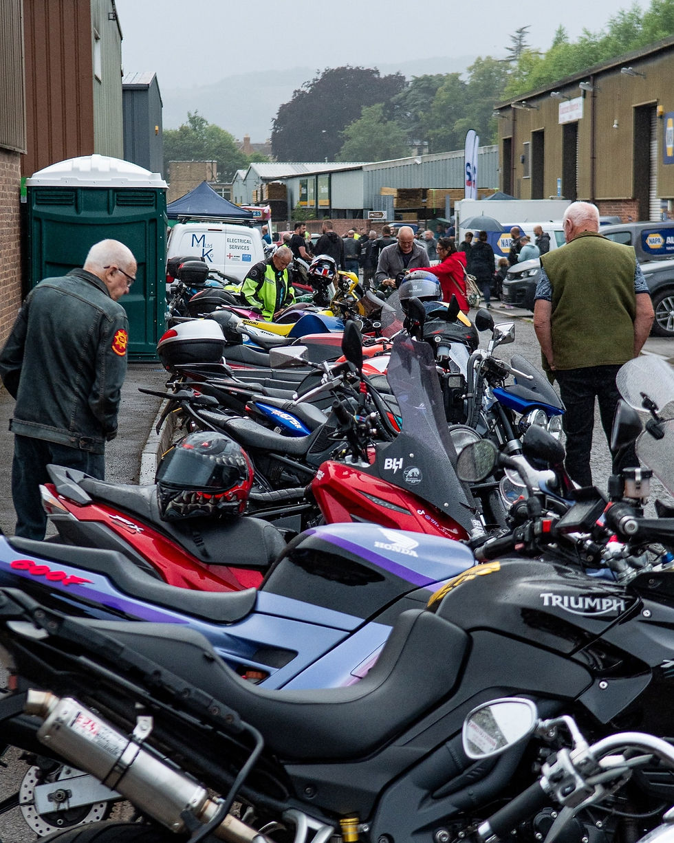 Motorcycles lined up on a street; people gather nearby, inspecting bikes. Overcast sky, industrial buildings, and trees in the background.
