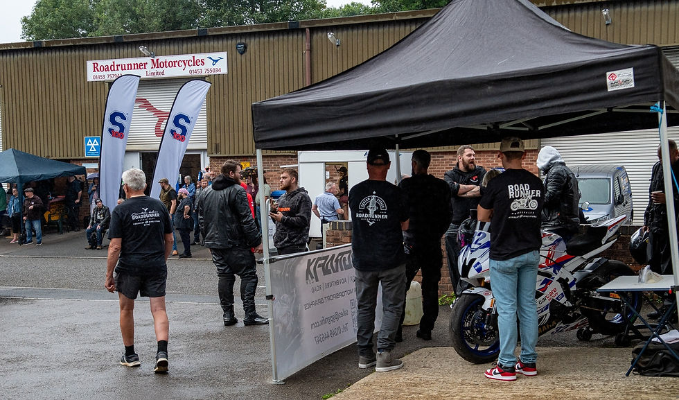 People at a motorcycle event gather near tents and a shop. Motorcycles and banners are visible. Casual atmosphere, cloudy day.