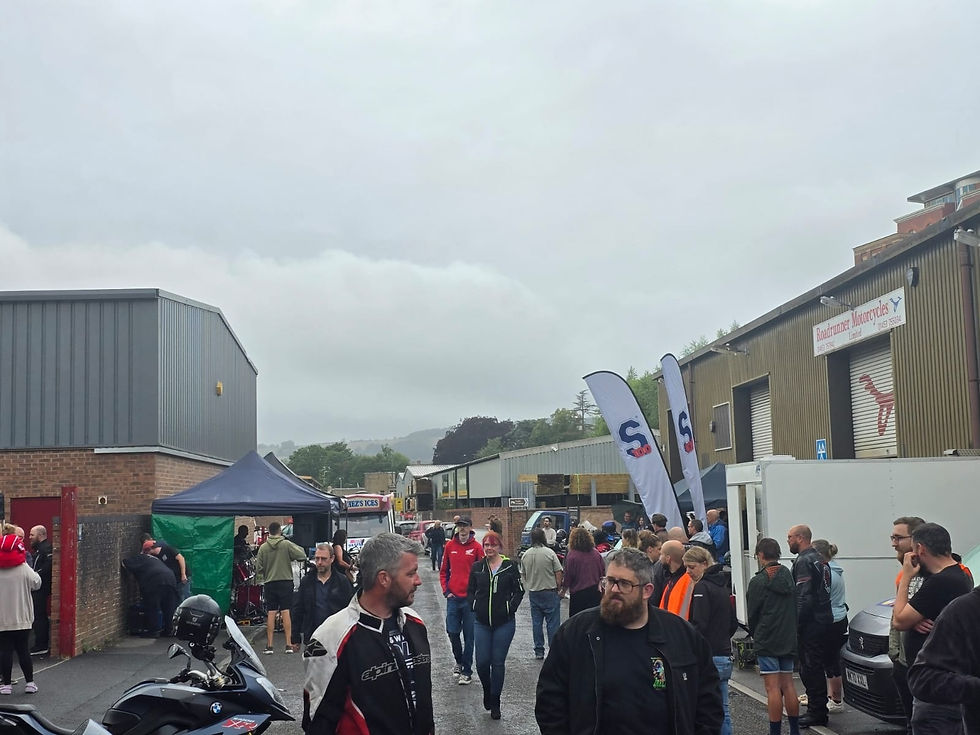 Crowd at motorcycle event outside industrial buildings. Overcast sky, banners, and motorcycle visible. People chatting and browsing.