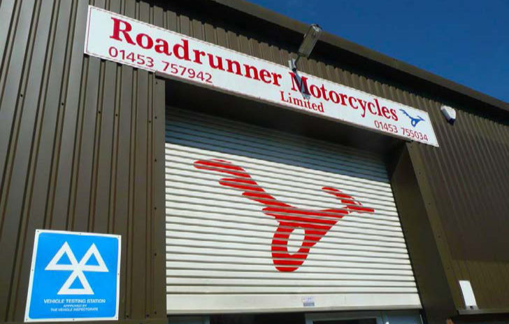 Roadrunner Motorcycles shop front with red logo on shutter and blue Vehicle Testing Station sign on a brown metal building.