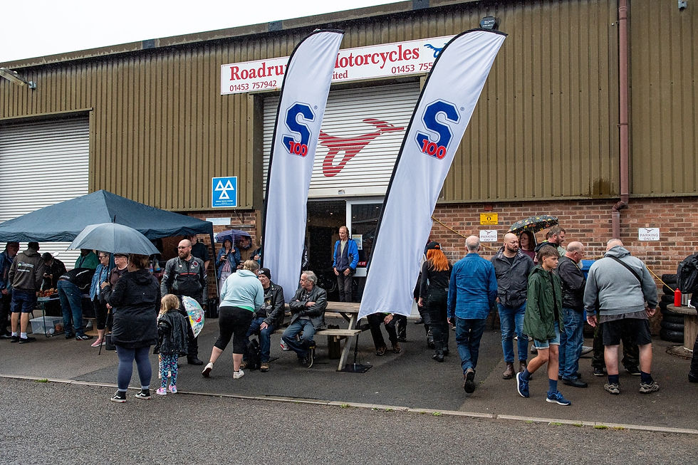Crowd outside a motorcycle shop with umbrellas in rain. Flags display "S 100." People in jackets, socializing, near a brick building.