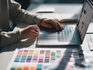 Person using a laptop on a desk with colour swatches. Sunlight casts shadows. Open notebook and pencil nearby, creating a focused mood.
