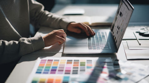 Person using a laptop on a desk with colour swatches. Sunlight casts shadows. Open notebook and pencil nearby, creating a focused mood.