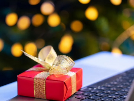 Small red gift box with a gold ribbon resting on a laptop keyboard, with holiday lights blurred in the background.