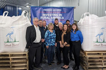 A group of nine people stand in front of a blue banner. There are large totes filled with grain on either side of the group.