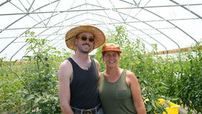 Two people stand in front of a hoop house with tomatoes.