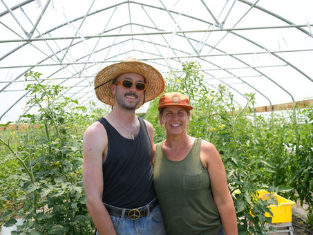 Two people stand in front of a hoop house with tomatoes.