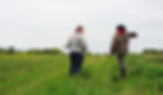 Two people stand in a field, both holding containers for planting.