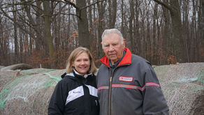 Two people stand in front of hay bails with a forest in the background.