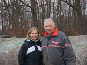 Two people stand in front of hay bails with a forest in the background. 