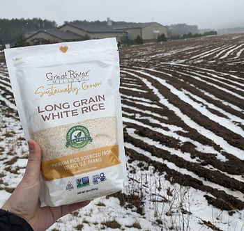 Someone holds a bag of rice. There is a farm field in the background with a dusting of snow.