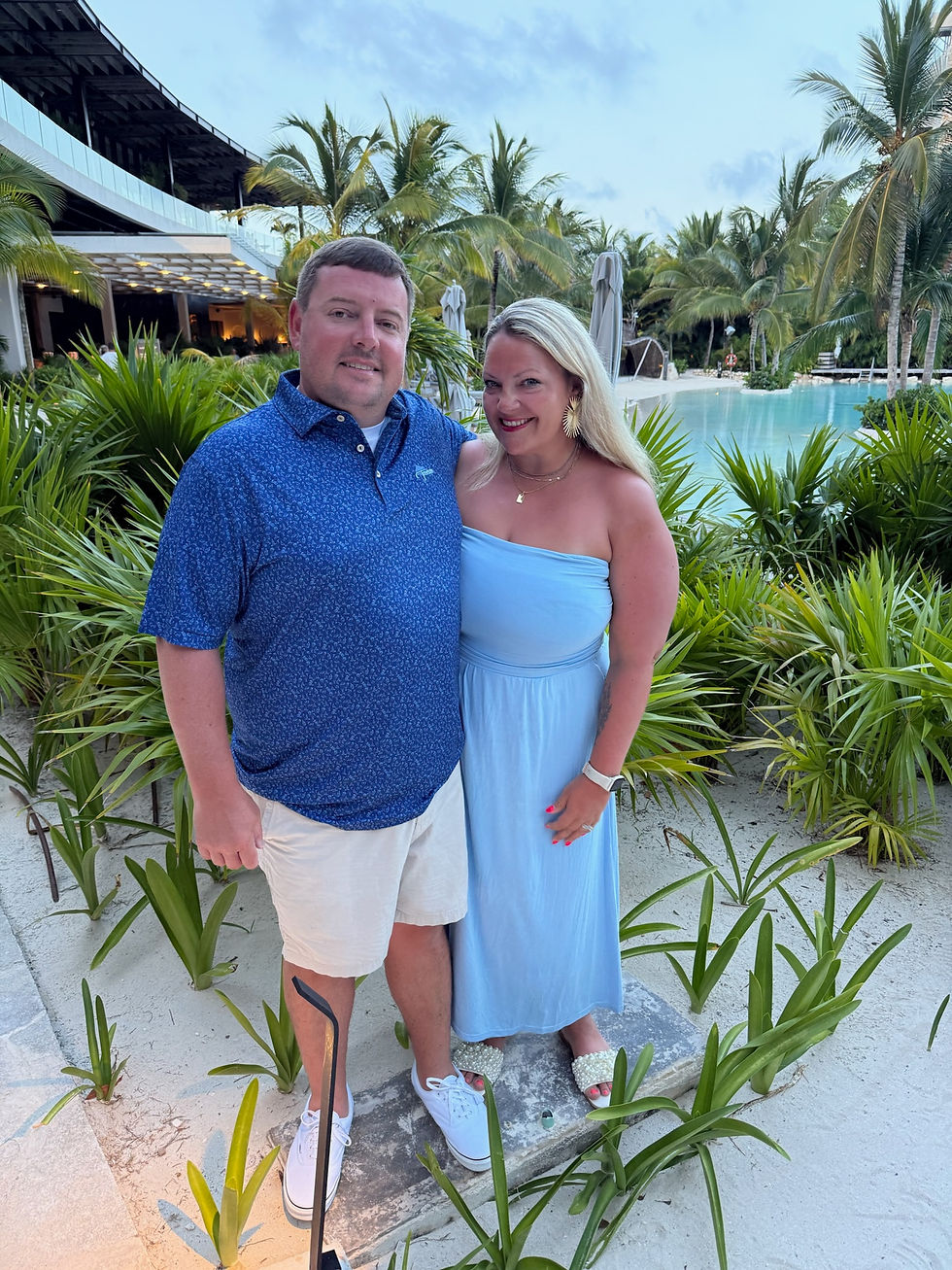 Smiling couple pose by a tropical resort pool, surrounded by palm trees and greenery, man in blue polo, woman in light blue dress.