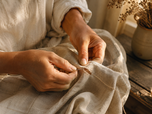 Hands sewing a tear in beige linen with needle and thread, in warm sunlight beside a rustic table and dried flowers.