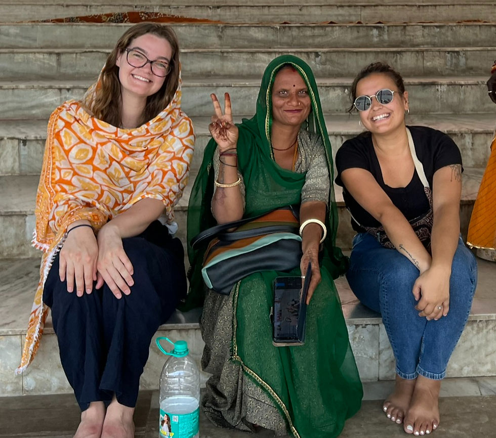 Three women sitting on steps, smiling. The woman in the middle wears green and gives a peace sign. A water bottle is on the ground., Pushkar, India