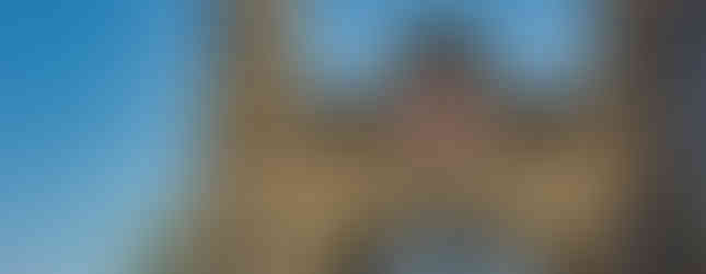 Sandsteinernes Brückentor mit zwei Türmen, Rundbogen und Wappen, dahinter sichtbare Metallkonstruktion der Brücke, umgeben von Bäumen und blauem Himmel.