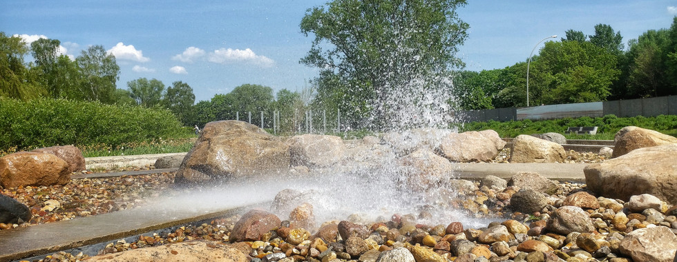 Wasserfontäne, die kraftvoll aus einem Natursteinbereich aufsteigt, umgeben von Steinen, Grünflächen und Bäumen unter blauem Himmel im Wilhelmsburger Inselpark.