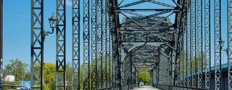 Stahlfachwerkbrücke mit offenen Dreiecksstrukturen, dekorativen Laternen und blauem Himmel, umgeben von Bäumen.