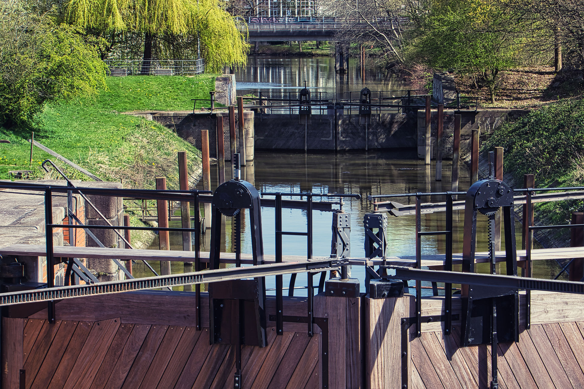 Geschlossene Schleuse mit Holz- und Metalltoren, gefülltem Schleusenbecken und umliegenden Grünflächen, Bäumen und Böschungen unter blauem Himmel.
