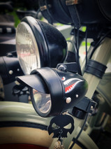 Nahaufnahme eines Custom‑Bike‑Scheinwerfers mit Hai‑Mund‑Design und kleinem Totenkopf‑Anhänger, montiert an einem Retro‑Motorrad‑Lenker.