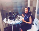 Mai Ling is seated in a podcast booth with a dark blue sleeveless dress on and a large pair of black headphones. On the round desk in front of her are a laptop and a number of stands with headphones. 